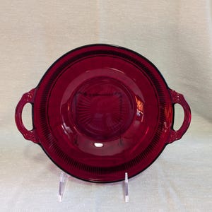 May include: A red glass serving dish with two handles. The dish has a raised rim and a textured bottom.