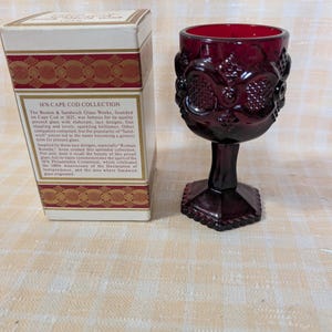 May include: A ruby red glass goblet with ornate floral and geometric patterns, next to a box with text that reads "1876 Cape Cod Collection". The goblet has a hexagonal base and a detailed bowl. The box has gold accents.