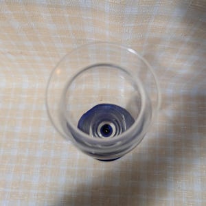 May include: A clear glass with a blue base, viewed from above. The glass has a rounded bowl and a stem that transitions to a dark blue color at the base. The background is a light-colored, checkered pattern.