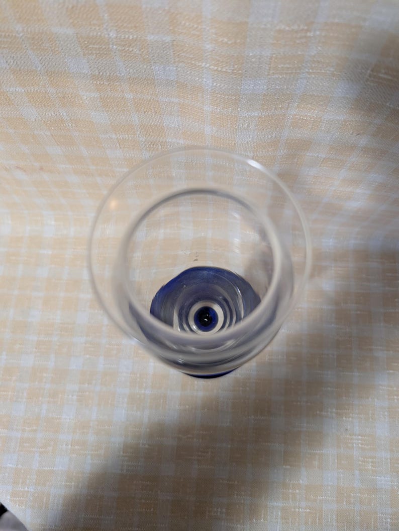 May include: A clear glass with a blue base, viewed from above. The glass has a rounded bowl and a stem that transitions to a dark blue color at the base. The background is a light-colored, checkered pattern.