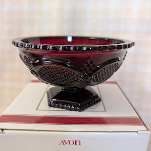 May include: A ruby-red glass bowl from the Avon 1876 Cape Cod Collection. The bowl has a decorative, textured pattern and sits on a square pedestal base. The rim of the bowl has a scalloped edge. The bowl is displayed on its original box.