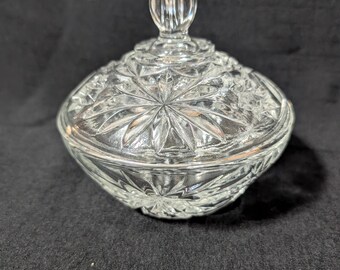 May include: A clear, cut-glass candy dish with a lid. The dish has a starburst pattern on the lid and bowl. The lid has a rounded, textured handle. The dish is sitting on a black surface.