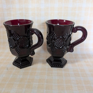 May include: Two ruby red glass mugs with ornate floral and geometric patterns. Each mug has a handle and sits on a hexagonal base. The mugs are likely vintage or antique and are displayed on a light-colored, patterned surface.