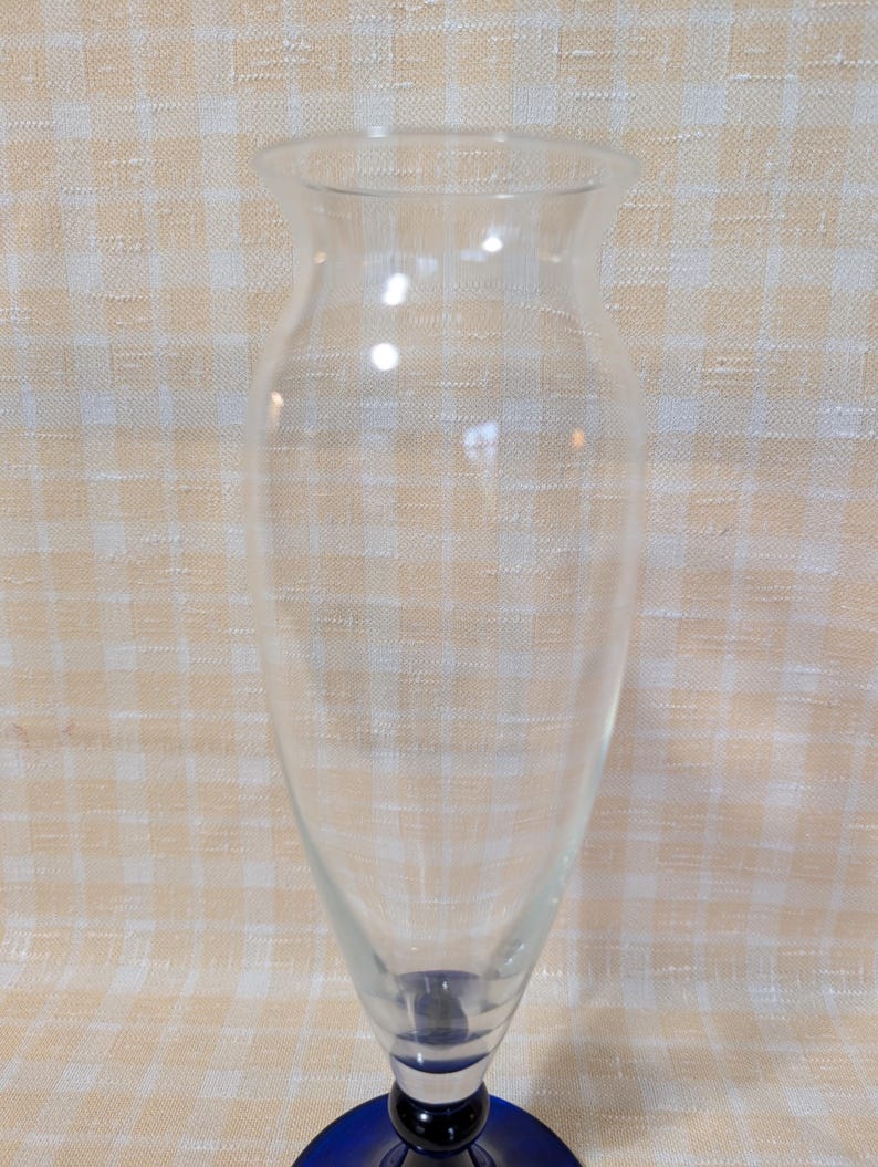 May include: A clear glass vase with a flared top and a blue base. The vase has a slender, elongated shape, perfect for displaying flowers or decorative branches. The blue base adds a pop of color to the otherwise transparent design.