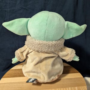 Puede incluir: Un juguete de peluche del personaje Baby Yoda de la serie de Star Wars The Mandalorian. El juguete lleva un abrigo marr&oacute;n con un cuello de piel sint&eacute;tica y est&aacute; sentado sobre una superficie de madera. El juguete es verde con orejas grandes y una boca peque&ntilde;a.