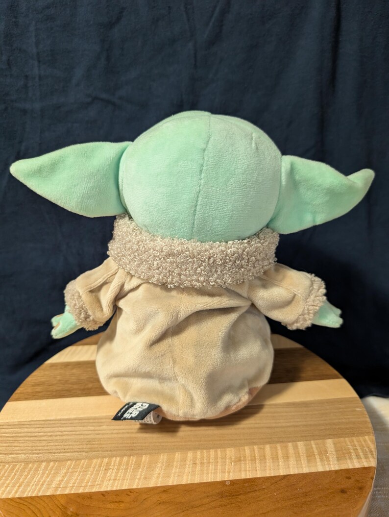 Puede incluir: Un juguete de peluche del personaje Baby Yoda de la serie de Star Wars The Mandalorian. El juguete lleva un abrigo marr&oacute;n con un cuello de piel sint&eacute;tica y est&aacute; sentado sobre una superficie de madera. El juguete es verde con orejas grandes y una boca peque&ntilde;a.