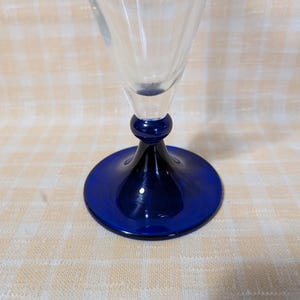 May include: A clear glass wine glass with a cobalt blue base and stem. The base is a flat circle, and the stem has a rounded bulb shape. The glass is empty and sits on a light-colored checkered surface.
