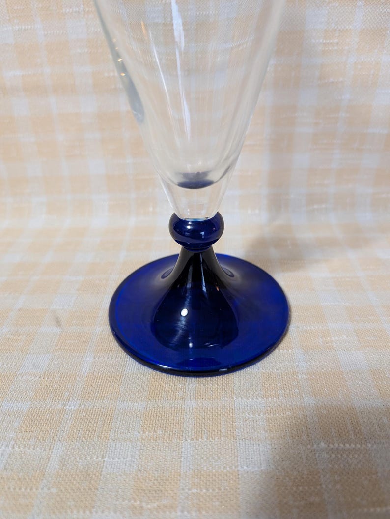May include: A clear glass wine glass with a cobalt blue base and stem. The base is a flat circle, and the stem has a rounded bulb shape. The glass is empty and sits on a light-colored checkered surface.