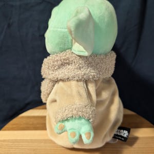 Puede incluir: Un juguete de peluche del personaje Baby Yoda de la serie Star Wars, que lleva una t&uacute;nica marr&oacute;n con capucha y ribete de piel. El juguete es verde con orejas grandes y est&aacute; de pie sobre una superficie de madera.