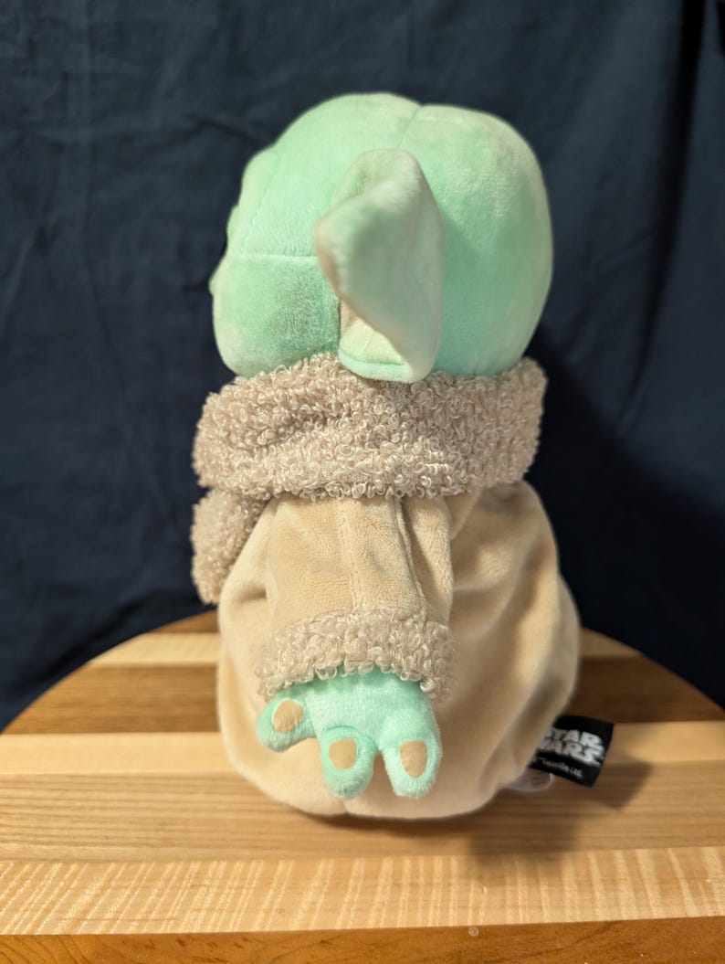 Puede incluir: Un juguete de peluche del personaje Baby Yoda de la serie Star Wars, que lleva una t&uacute;nica marr&oacute;n con capucha y ribete de piel. El juguete es verde con orejas grandes y est&aacute; de pie sobre una superficie de madera.