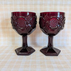 May include: Two ruby red goblets with ornate, embossed designs. The glasses have a detailed pattern around the bowl and a faceted base. The stemware is made of a deep red glass, perfect for serving beverages.