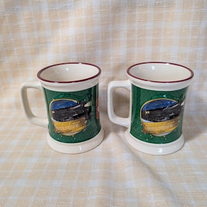 May include: Two ceramic mugs featuring a green background with a dark green diamond pattern. Each mug displays a graphic of a black steam train under a starry night sky with the text The Polar Express and the word BELIEVE in gold lettering.