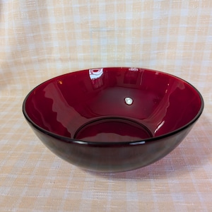 May include: A deep red glass bowl with a glossy finish. The bowl has a rounded shape with a dark rim. The interior reflects light, and the bowl sits on a light-colored, checkered surface.