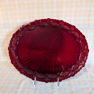 May include: A ruby red oval serving platter with a decorative scalloped edge. The platter has a textured, radial design in the center and a repeating pattern around the rim. The platter is displayed on a clear stand.