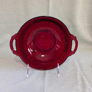 May include: A red glass serving dish with two handles. The dish has a fluted edge and a raised center.