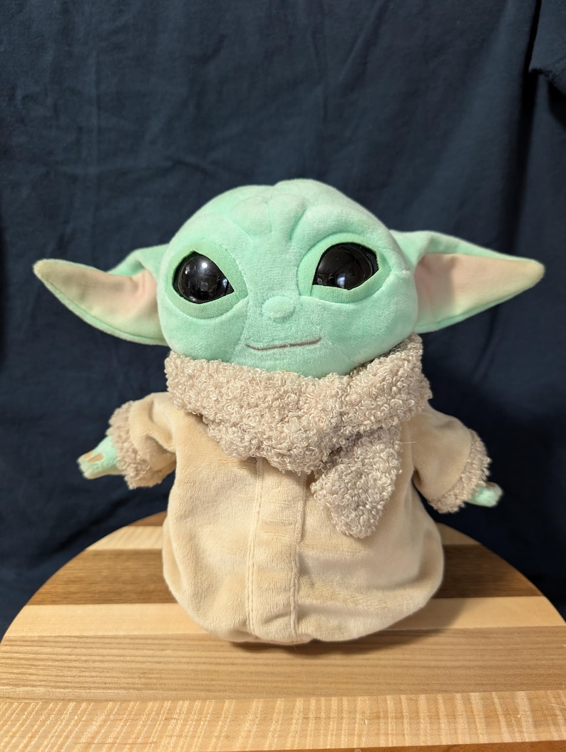 Puede incluir: Un juguete de peluche verde de un personaje de la franquicia Star Wars, conocido como "The Child" o "Baby Yoda". El juguete lleva un abrigo de peluche marr&oacute;n con una bufanda de piel marr&oacute;n.