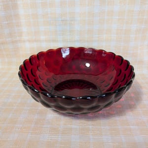May include: A ruby red glass bowl with a scalloped edge and a textured, bubble-like pattern. The bowl is round and sits on a light-colored, checkered surface. The bowl is empty.