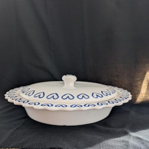 May include: White ceramic serving dish with a lid. The dish has a scalloped edge and is decorated with blue heart-shaped designs. The lid has a small, round handle. The dish is on a dark gray surface.