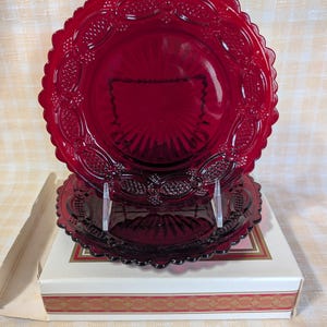 May include: Two ruby red glass plates with a scalloped edge and embossed grape design. The plates are stacked on a white box with a gold and red decorative border. The plates are supported by clear plastic stands.