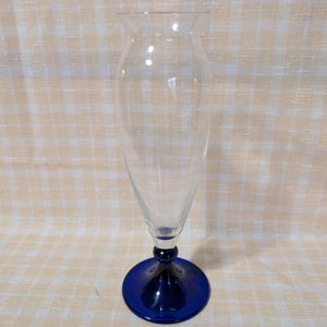 May include: A clear glass vase with a flared top and a conical body. The base is a deep blue, circular foot. The vase is empty and sits on a light-colored, checkered surface. The vase is designed for displaying flowers.
