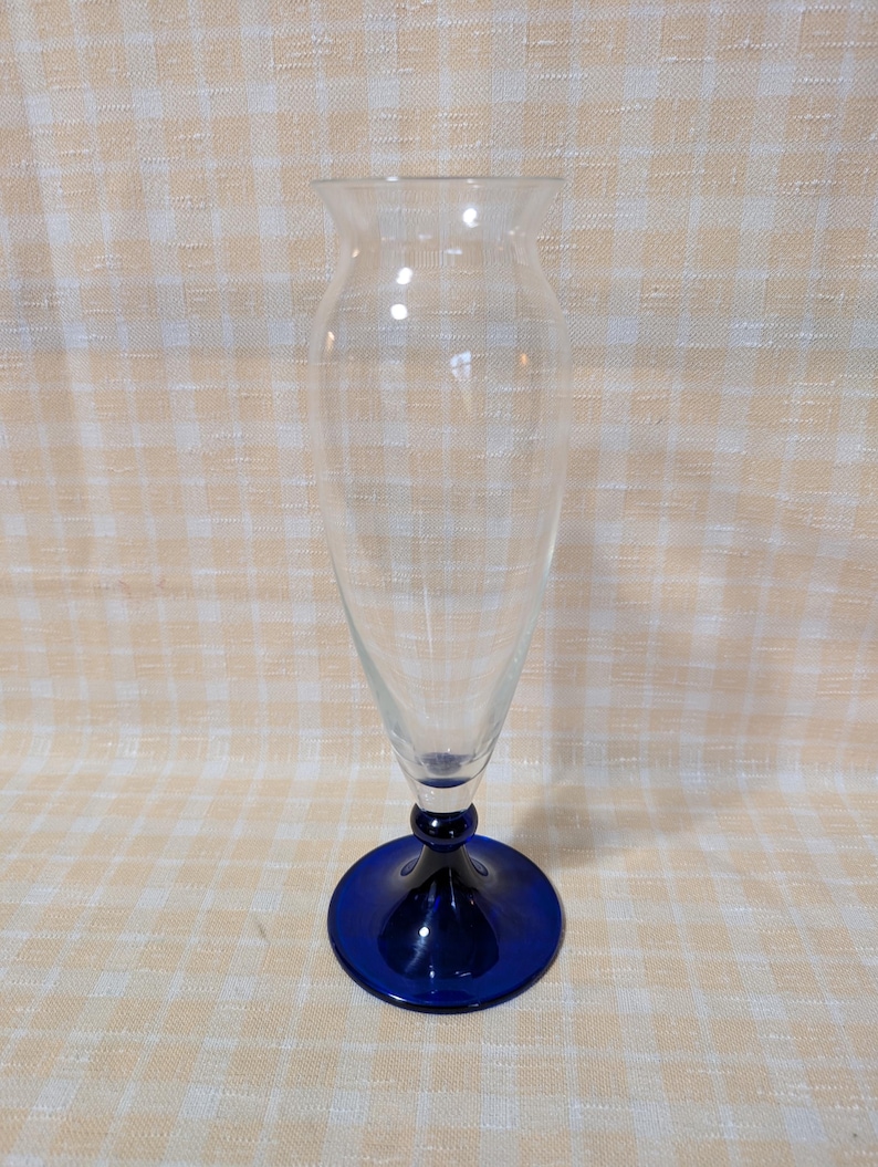 May include: A clear glass vase with a flared top and a conical body. The base is a deep blue, circular foot. The vase is empty and sits on a light-colored, checkered surface. The vase is designed for displaying flowers.