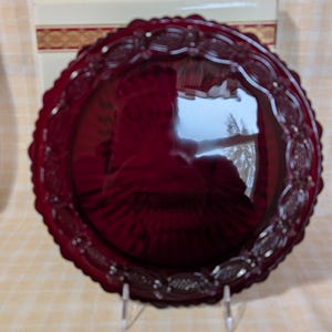May include: A ruby red glass plate with a scalloped edge and embossed floral design. The plate is displayed on a clear stand, with a box in the background. The plate has a reflective surface.