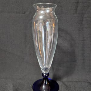 May include: A clear glass vase with a flared top and a slender, ribbed body. The vase sits on a dark blue, circular base. The stem has a bulbous shape. The vase is designed for floral arrangements.