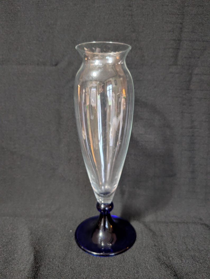 May include: A clear glass vase with a flared top and a slender, ribbed body. The vase sits on a dark blue, circular base. The stem has a bulbous shape. The vase is designed for floral arrangements.