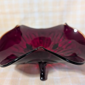 May include: A ruby red glass dish with a unique, petal-like shape. The dish has a dark, almost black, stem and a gold rim. The glass is transparent, allowing light to pass through, and is likely vintage.