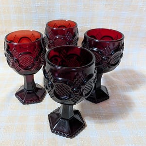May include: Four ruby red vintage goblets with ornate, textured designs. Each goblet features a hexagonal base, a stem, and a round bowl. The goblets are arranged on a light-colored surface, showcasing their rich color and intricate details.