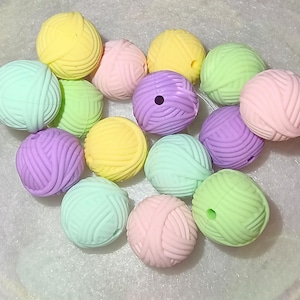 Op de afbeelding: Een verzameling kleurrijke, pastelkleurige garenbolkralen. De kralen zijn in tinten geel, roze, groen en paars. Elke kraal is rond met een gestructureerd oppervlak, dat lijkt op een bol garen. Geschikt voor knutselprojecten.