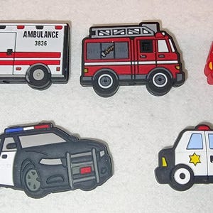 May include: A collection of colorful emergency vehicle-themed charms. Includes an ambulance with "AMBULANCE 3836" and "701" text, a fire truck, a fire engine, a police car, and a police car with "POLICE" text.