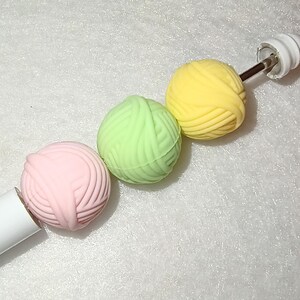 Op de afbeelding: Een witte breinaald met drie kleurrijke garenballen. De garenballen zijn roze, groen en geel. De naald heeft een witte handgreep en een witte eindkap.