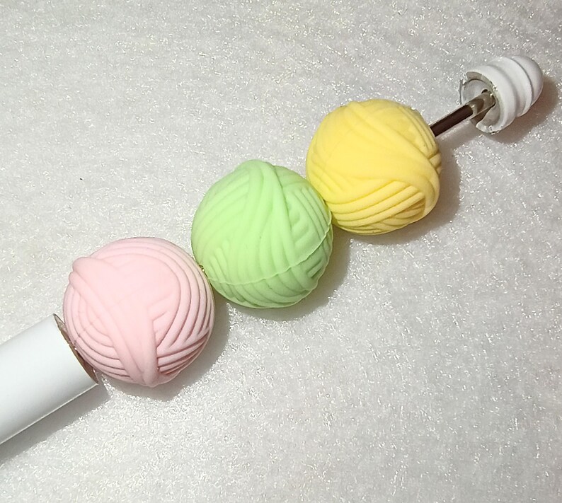 Op de afbeelding: Een witte breinaald met drie kleurrijke garenballen. De garenballen zijn roze, groen en geel. De naald heeft een witte handgreep en een witte eindkap.