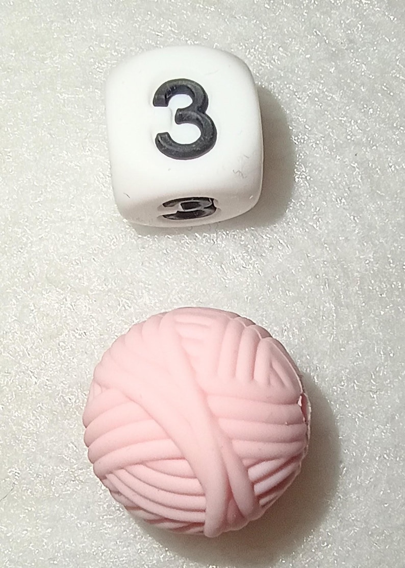 Op de afbeelding: Een witte kubus met het zwarte nummer 3 en een roze bol wol. De kubus heeft een klein gaatje aan de onderkant. De bol wol is lichtroze en heeft een getextureerd oppervlak.