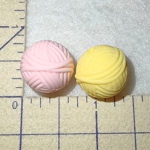 Op de afbeelding: Twee kleine, ronde gummen in de vorm van bollen wol, &eacute;&eacute;n lichtroze en de andere geel. Ze liggen op een licht oppervlak naast een liniaal, wat wijst op een knutsel- of kantoorartikelen thema. De gummen zijn ongeveer 1 cm in diameter.