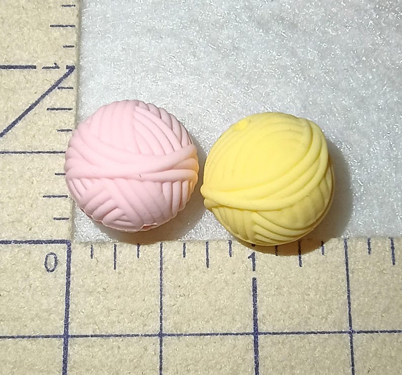 Op de afbeelding: Twee kleine, ronde gummen in de vorm van bollen wol, &eacute;&eacute;n lichtroze en de andere geel. Ze liggen op een licht oppervlak naast een liniaal, wat wijst op een knutsel- of kantoorartikelen thema. De gummen zijn ongeveer 1 cm in diameter.