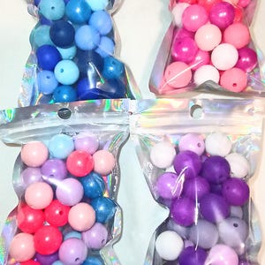 May include: Four clear plastic bags filled with colorful round beads. The beads are in shades of blue, pink, purple, and white. The bags have a holographic, iridescent sheen.