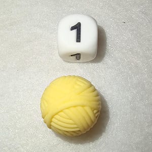 Op de afbeelding: Een witte dobbelsteen met het zwarte nummer 1 en een gele bol wol. De dobbelsteen is kubusvormig en de wol is een strak opgerolde bol. De objecten staan tegen een witte, getextureerde achtergrond.