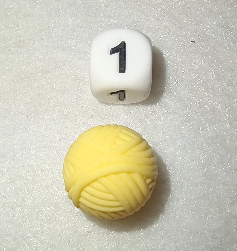 Op de afbeelding: Een witte dobbelsteen met het zwarte nummer 1 en een gele bol wol. De dobbelsteen is kubusvormig en de wol is een strak opgerolde bol. De objecten staan tegen een witte, getextureerde achtergrond.
