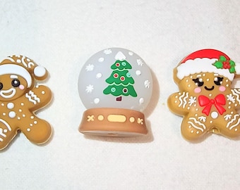 May include: Three Christmas-themed decorations: two gingerbread men and a snow globe. The gingerbread men are brown with white icing details and festive hats. The snow globe features a green Christmas tree and snowflakes.