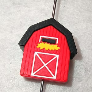 May include: A red barn-shaped bead with a black roof and white door design. The bead has a yellow hay detail and is strung on a silver-colored wire. This farm-themed bead is suitable for jewelry making.