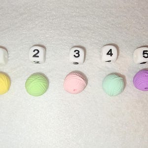 Op de afbeelding: Een bovenaanzicht toont vijf witte dobbelstenen met zwarte cijfers van &eacute;&eacute;n tot vijf, elk gecombineerd met een kleine bol wol in bijpassende kleuren: geel, groen, roze, mint en paars. De items zijn in een rij gerangschikt.
