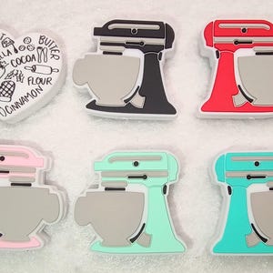 May include: A collection of colorful stand mixer-shaped charms in black, red, pink, mint green, and teal. A white heart-shaped charm with baking-related text and illustrations is also included. These charms are likely for crafting or decorating.