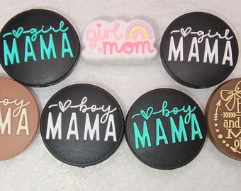 Op de afbeelding: Een verzameling van zeven ronde en één wolkvormige siliconen bedels. De bedels bevatten teksten zoals girl MAMA, girl mom, boy MAMA en bows and ARROWS MOM OF BOTH. De ontwerpen zijn in zwart, bruin, wit, turquoise en roze met hartjes.