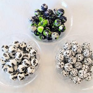 May include: Three bowls of colorful silicone beads. The first bowl contains black, white, and green beads. The second bowl contains black and white beads with a paw print design. The third bowl contains black and white beads with a musical note design.