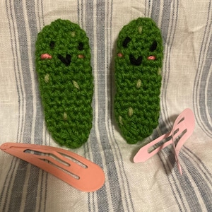May include: Two green crochet pickle hair clips with black eyes, smiles, and pink cheeks. Two pink snap hair clips are also present. The items are on a striped fabric background.