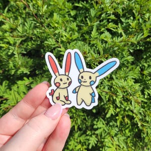 May include: Two cartoon stickers of a yellow Pokemon character with red and blue accents. The character on the left has red ears and a red heart on its chest. The character on the right has blue ears and a blue heart on its chest.