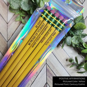 May include: A pack of yellow pencils with pink erasers, each printed with a positive affirmation in black. The pencils are contained in a clear, iridescent cellophane bag. Affirmations include "BE KIND" and "YOU GOT THIS!"