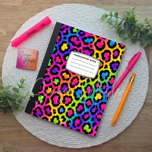 May include: A composition book with a vibrant rainbow leopard print pattern, a black spine, and a white label featuring the text COMPOSITION BOOK. Included are a pink Sharpie highlighter, a pink pen, an orange pencil, and a small square tag.